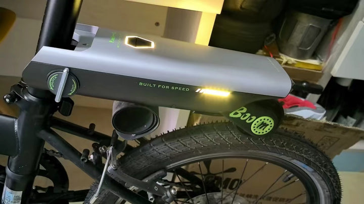 A close-up of a bicycle battery mounted on a black bicycle frame in a cluttered indoor setting. The battery is silver and black with green accents, including the brand name &