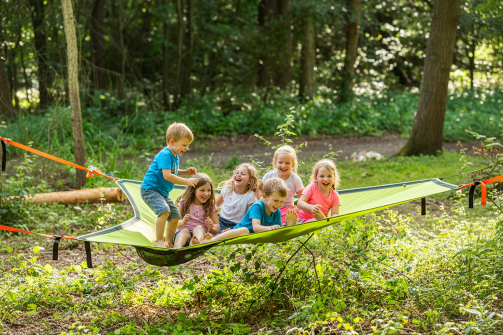 A group of five children playing in a bright green camping hammock suspended between trees in a wooded outdoor setting.